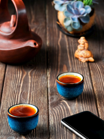 两个碗与红色的茶粘土茶壶的表格茶仪式智能手机的表格下一个的碗新鲜酿造黑色的茶蒸汽上升以上的杯黑暗木背景特写镜头