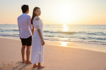 年轻的快乐亚洲夫妇持有手的海滩与女人面对相机鼓浪漫的旅行度蜜月夏天假期亚洲女人和男人。持有手拥抱在户外日落背景