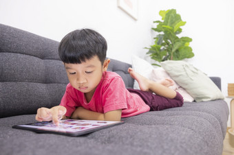微笑小男孩看起来的触控板说谎舒适的沙发的生活房间享受玩游戏数字平板电脑快乐小孩子使用有趣的应用程序