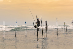 高跷渔夫坐着他的波兰与木钓鱼杆他的手的日落晚上海洋波崩溃后面他的背景