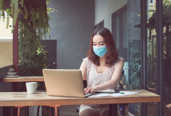 亚洲美丽的女人穿面具保护病毒和使用移动PC为工作首页新正常的和技术概念