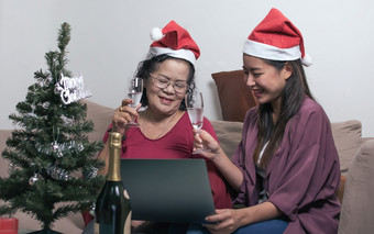 两个老妈妈。和女儿穿圣诞老人他喝和在线闲谈，聊天与其他人使用移动PC而坐着生活房间首页与模糊前景树和瓶圣诞节新一年庆祝活动概念