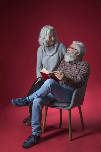<strong>高级</strong>女人坐着与她的丈夫坐着椅子持有书手对<strong>红色</strong>的<strong>背景</strong>