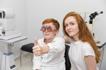 男孩与验光师试验框架显示拇指手势坐着与年轻的女<strong>眼科</strong>医生<strong>诊所</strong>