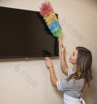 微笑肖像年轻的女人使用除尘器清洁电视屏幕墙