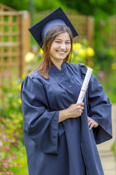微笑年轻的女人持有文凭和穿帽和礼服在户外看相机毕业概念