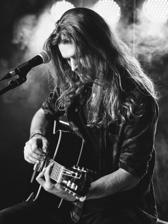 照片年轻的男人。与长头发和胡子唱歌和玩声吉他阶段与灯和音乐会大气