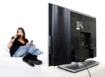 年轻的女人坐着舒适的白色沙发上喝杯茶与远程控制她的手看巨大的平屏幕电视