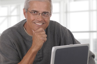 微笑随便穿着成熟的商人坐着后面移动PC电脑与他的手他的下巴的男人。前面大现代办公室窗口