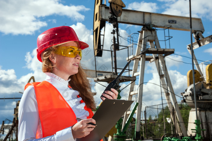 女人工程师的石油场会说话的的广播穿红色的头盔和橙色工作衣服工业网站背景石油和气体概念