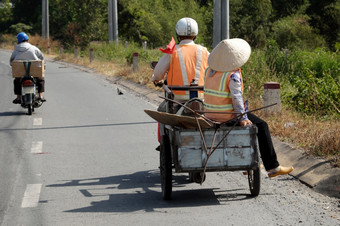 两个环境卫生工人移动国家路在一起男人。骑摩托车与拖盒子转移女人夫妇的道路后完成工作他们的影子反映表面路越南