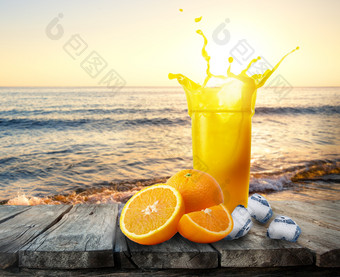 玻璃橙色汁与飞溅橙子和冰木表格玻璃汁站表格在的海日落的概念休息和假期玻璃橙色汁与飞溅橙子和冰木