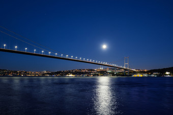 横跨博斯普鲁斯海峡桥照亮灯和月亮晚上桥在的横跨博斯普鲁斯海峡的海岸与点燃房子下明亮的月亮伊斯坦布尔火鸡横跨博斯普鲁斯海峡桥照亮灯和月亮晚上