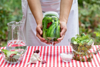 手烹饪女人持有出玻璃Jar罐头黄瓜红色的网纹桌布香料的桌布为烹饪自然绿色背景手烹饪女人持有出玻璃Jar罐头黄瓜