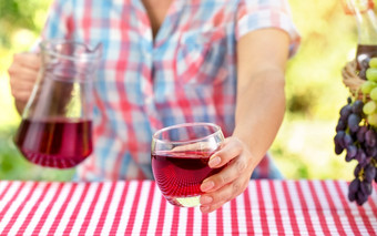 女人持有出玻璃酒葡萄汁在表格与红色的桌布附近的篮子葡萄手壶自然绿色背景野餐概念女人持有出玻璃酒葡萄汁在表格与红色的桌布