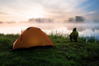 渔夫坐着海岸多雾的湖附近橙色帐篷早期早....渔夫坐着海岸多雾的湖附近橙色帐篷