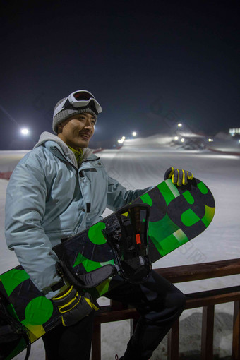 夜晚青年男人抱着滑雪板坐在滑雪场内的护栏上