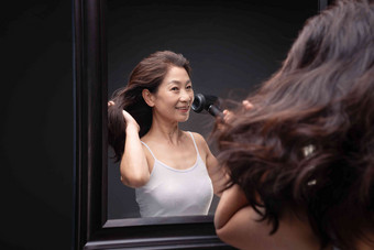 <strong>中年女人</strong>对着镜子用吹风机吹头发微笑高质量摄影