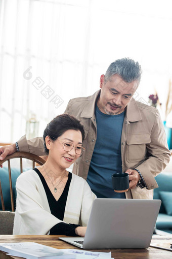 中年男士拿着杯子看使用笔记本电脑的妻子