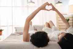 快乐的年轻情侣躺在床上做心形手势室内清晰图片
