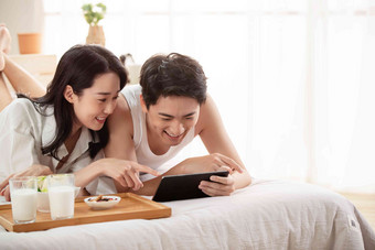 年轻情侣在床上一边吃早餐一边看平板电脑