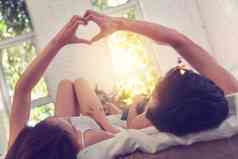 快乐的年轻情侣躺在床上做心形手势女人高质量图片