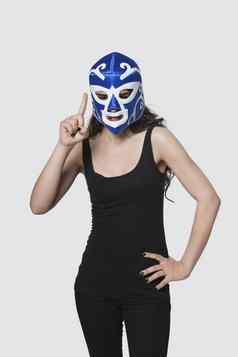 年轻的女穿摔跤面具提出了指数手指灰色的背景