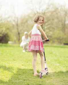 年轻的女孩骑踏板车公园妈妈。背景