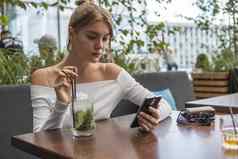 快乐的金发碧眼的女人博主鸡尾酒阅读传入的短信消息智能手机连接免费的无线网络咖啡馆积极的年轻的学生看广播在线现代移动电话