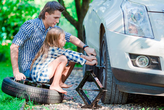 可爱的女孩坐着轮胎帮助父亲改变车轮在户外美丽的夏天一天