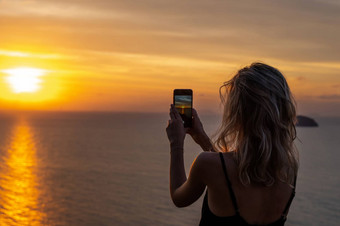 女人手持有移动电话日落年轻的卷曲的头发女人采取照片细胞电话美丽的令人惊异的日落海采取图片智能手机假期旅行