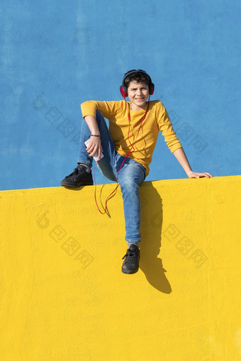 前面视图年轻的男孩穿休闲衣服坐着黄色的栅栏蓝色的墙移动电话听音乐耳机