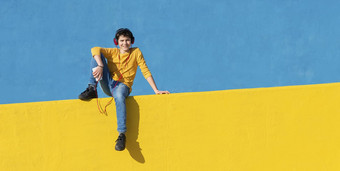 前面视图年轻的男孩穿休闲衣服坐着黄色的栅栏蓝色的墙移动电话听音乐耳机