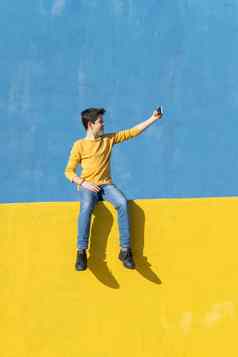 前面视图年轻的男孩穿休闲衣服坐着黄色的栅栏蓝色的墙采取自拍智能手机
