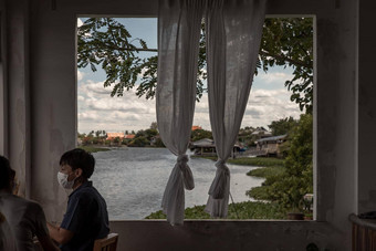 亚洲年轻的男人。穿脸面具坐着窗口框架白色花边窗帘美丽的河视图