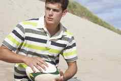 十几岁的男孩坐着海滩持有橄榄球球