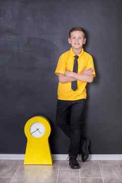 可爱的英俊的学校男孩黄色的t恤领带时尚的靴子casuals站关闭黑色的董事会黄色的时钟看微笑