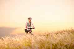年轻的女人骑山自行车美丽的场羽毛草日落冒险旅行