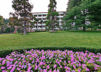 花圃软粉红色的紫色的花前面白色建筑酒店娱乐中心疗养院阳台背景高冷杉树绿色草坪上清晰的天空