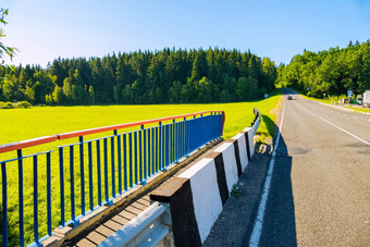 结束桥沥青路车通过沐浴在阳光里的长满草的草地密集的绿色松柏科的森林蓝色的万里无云的天空假期旅行
