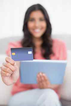 快乐可爱的浅黑肤色的女人坐着沙发上显示信贷卡持有平板电脑