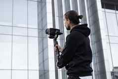 年轻的专业电视录像制作人持有专业相机轴健身球稳定剂为设备帮助使高质量视频摇晃摄影师穿白色连帽衫使视频