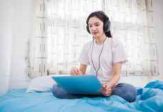 亚洲快乐年轻的女人穿耳机听音乐首歌看网站购物在线移动PC床上首页