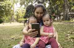 亚洲妈妈。女儿坐着草地面花园智能手机幸福的
