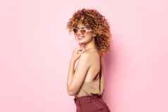 有吸引力的女人卷曲的头发微笑太阳镜粉红色的背景时尚衣服