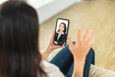 年轻的女人坐着沙发首页持有移动电话使视频调用讨论公司的同事们远程办公
