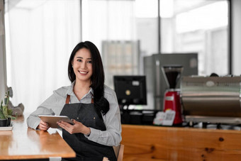 快乐的年轻的亚洲女人老板持有数字平板电脑站咖啡馆年轻的企业家概念上的