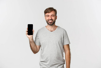 肖像微笑英俊的男人。灰色的t恤显示移动电话屏幕推荐应用程序<strong>购物网站</strong>站白色背景