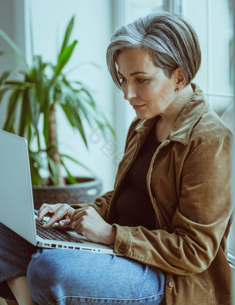 优雅的成熟的女人作品移动PC坐着窗台上窗口一边视图现代银头发的夫人休闲打字电脑键盘
