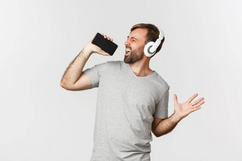 肖像英俊的的家伙灰色的t恤玩<strong>卡拉ok</strong>应用程序耳机唱歌移动电话麦克风站白色背景